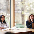 Two women sat at a desk with a notebook and a laptop listening to a speaker