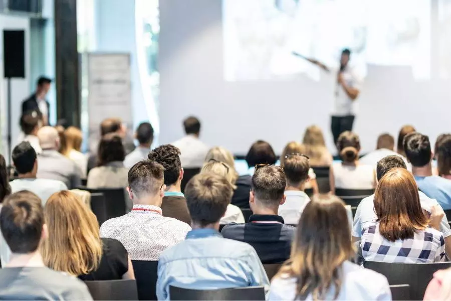 A large group of people sat down listening to someone on a stage talk into a microphone